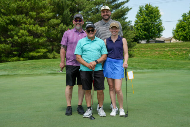 Three men and one woman are smiling on the green for a group photo.