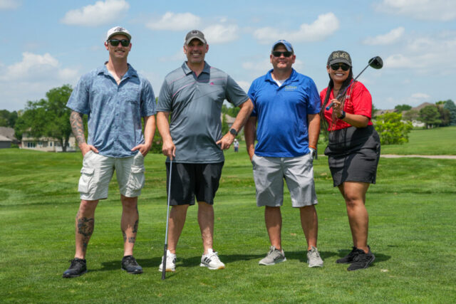 Three men and one woman are smiling on the green for a group photo.