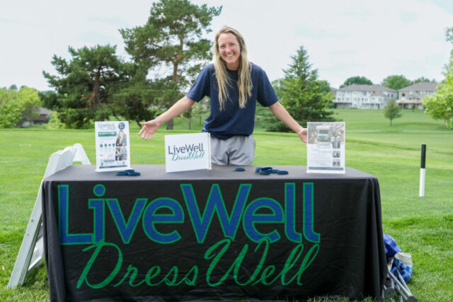A lady from LiveWell DressWell smiling behind her hole table.