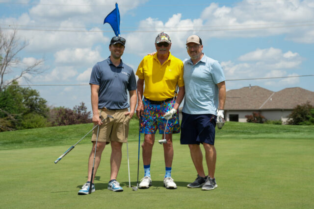 Three gentlemen are smiling on the green for a group photo.