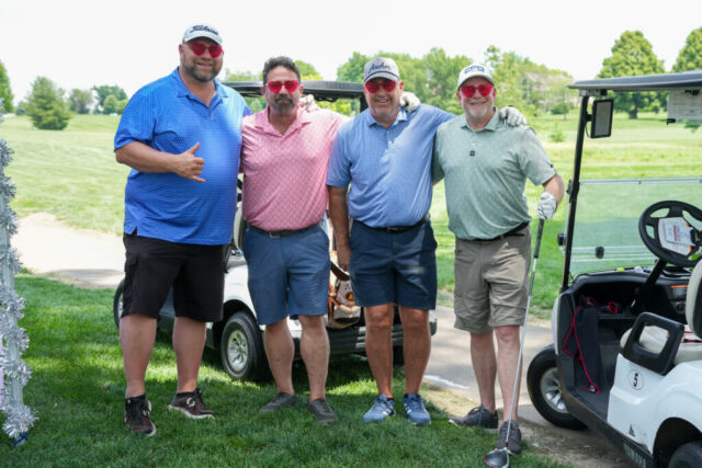 Four gentlemen wearing red heart sunglasses are smiling on the green for a group photo.