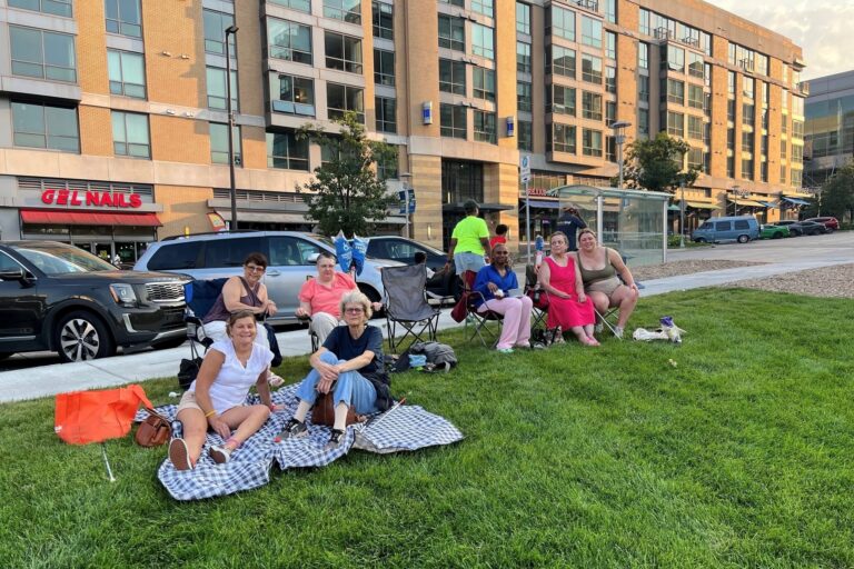 A group of ladies sitting on chairs and blankets in the grass at turner park. buildings of midtown crossing are in the background.