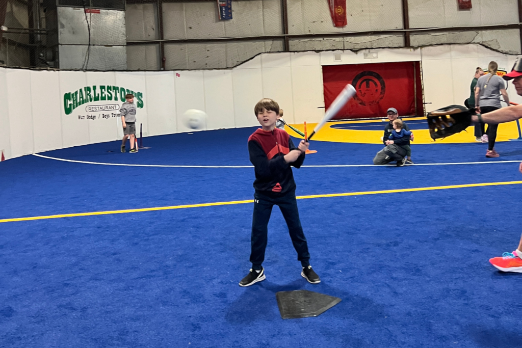 Reuben is at an indoor rec center, swinging at a baseball mid-air while a catcher is behind him.