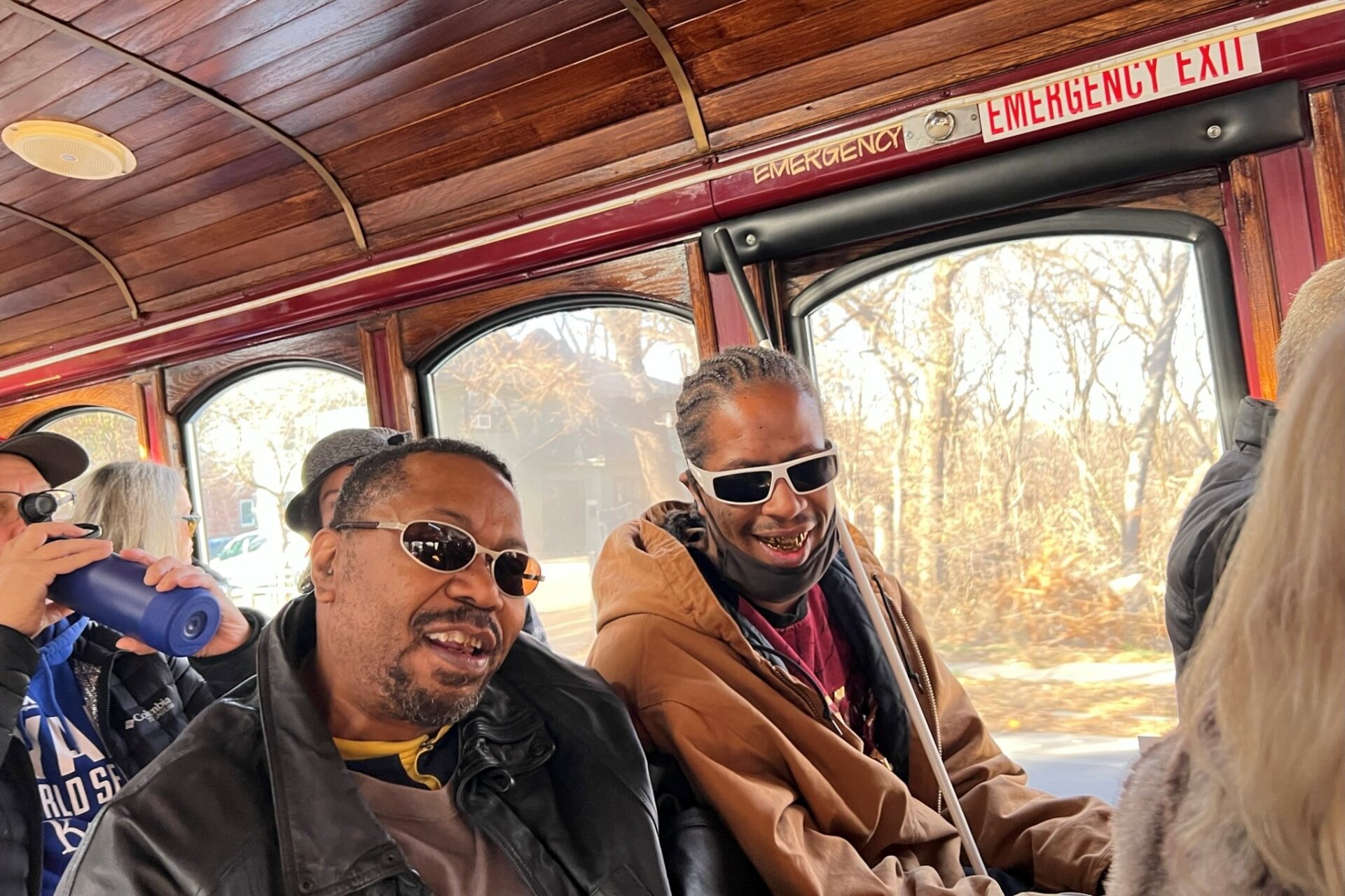 Two men sharing a bench on an Ollie the Trolley tour. They are smiling, one has a white cane and near a wind0w.