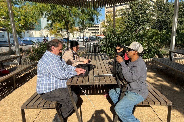 A small group of adults sitting outside downtown at a picnic table using computers and phones to write poems and short stories.
