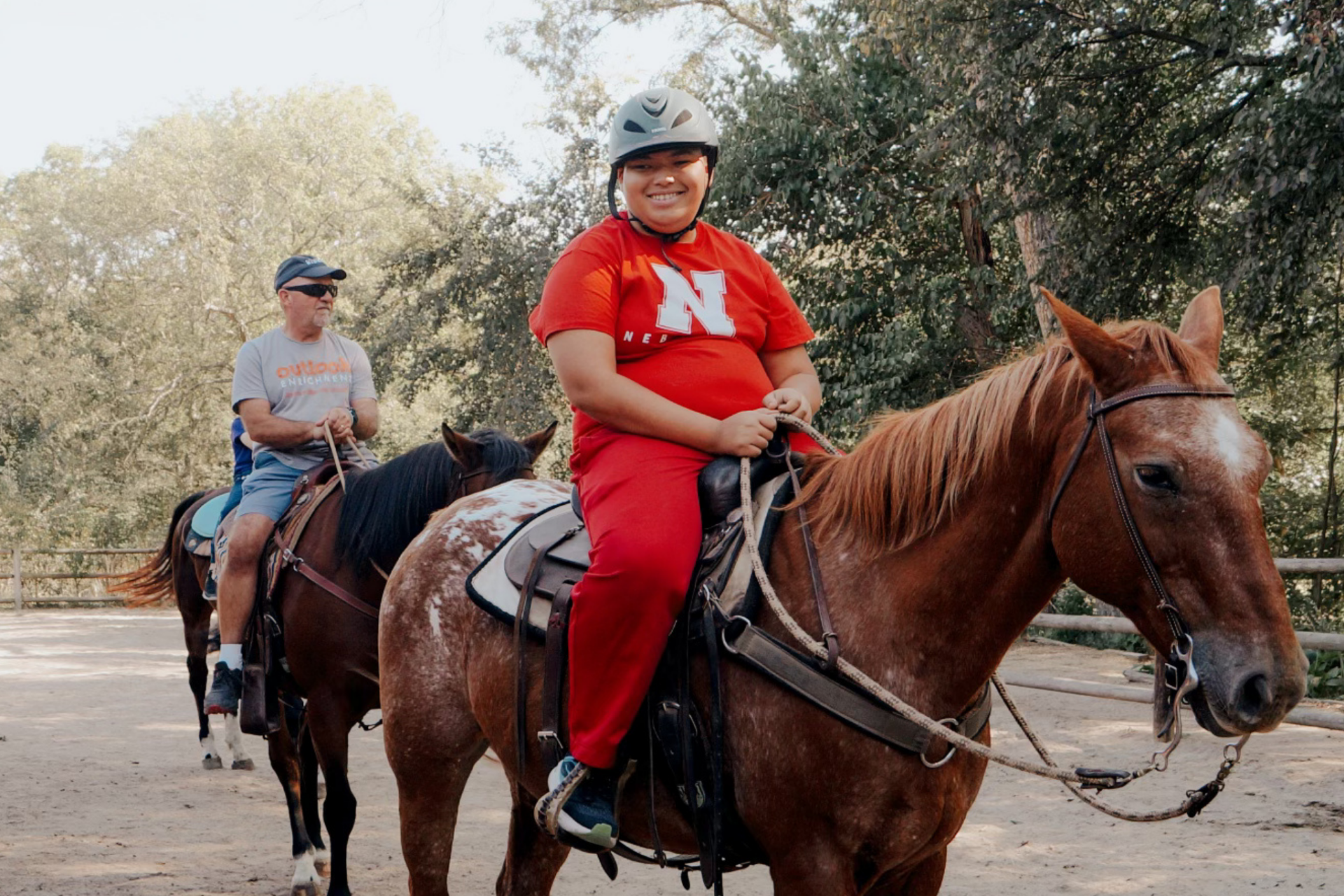 A young man on a brown horse wearing a riding helmet and a red Nebraska T-shirt and smiling.