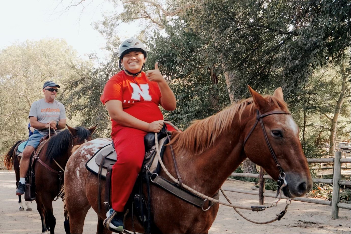 A young man on a brown horse wearing a riding helmet and a red nebraska t-shirt and smiling.