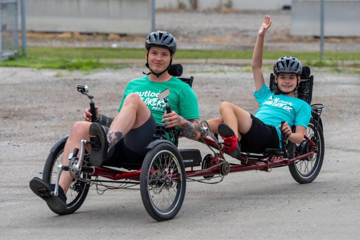 A young male participant waves while seated on the back of a tandem bike. While a male counselor, the bike captain, is seated and smiling in the front.